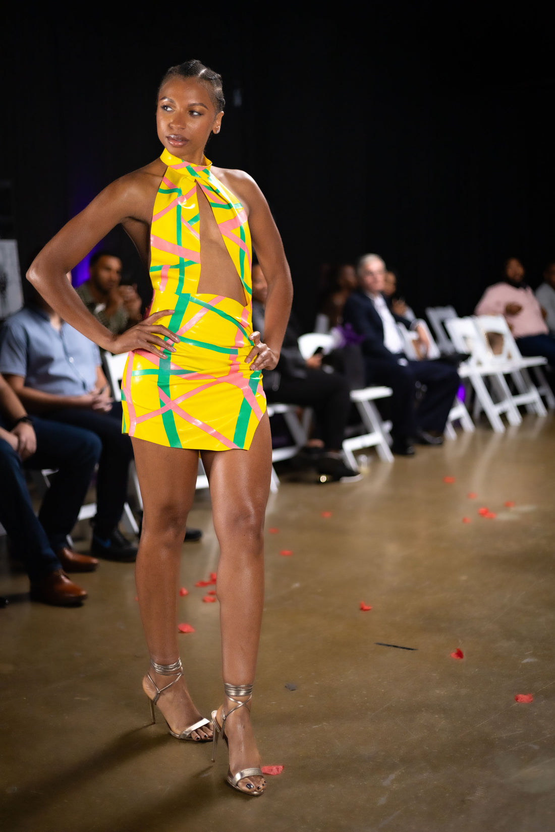 Model wearing a vibrant yellow and green dress on a runway with an audience in the background.