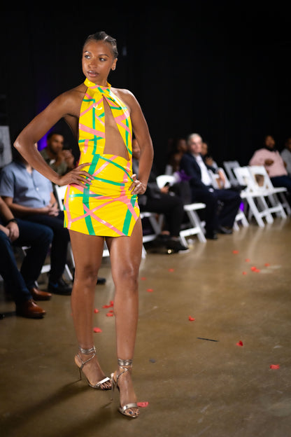 Model wearing a vibrant yellow and green dress on a runway with an audience in the background.