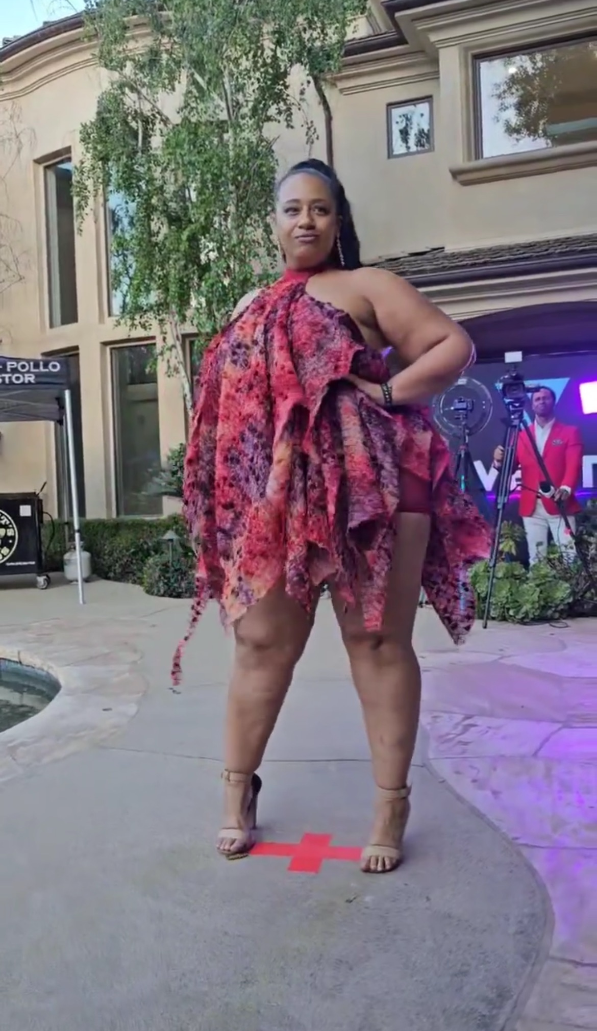 Woman in a colorful dress standing on a sidewalk with buildings and a TV screen in the background.