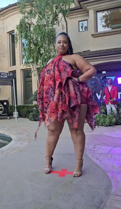 Woman in a colorful dress standing on a sidewalk with buildings and a TV screen in the background.