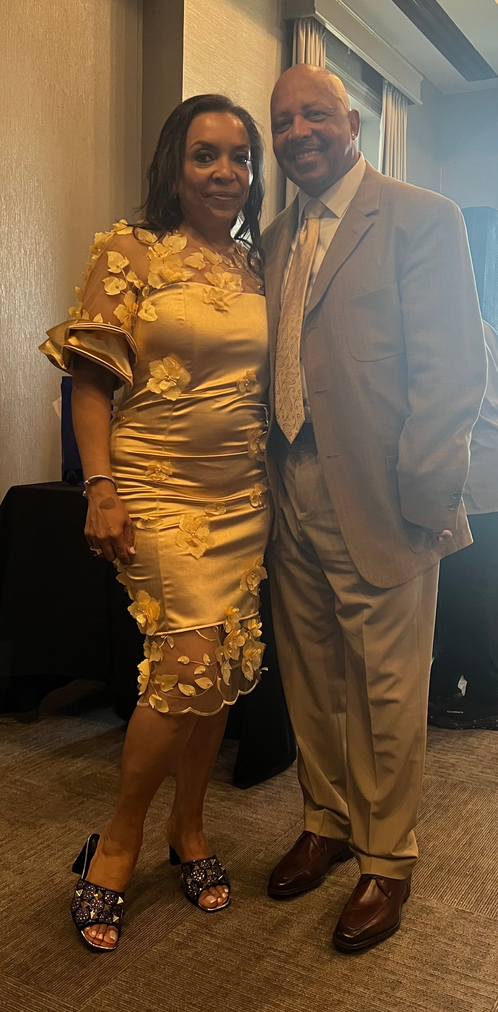 Man and woman posing together in a formal setting with chairs and a window in the background.
